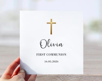 May include: A white card with a gold cross at the top. The name "Olivia" is written in cursive below the cross, followed by "FIRST COMMUNION" and the date "16.05.2026".