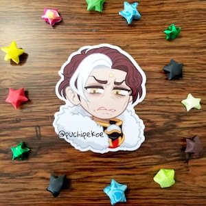 May include: A sticker depicting a cartoon character with white and burgundy hair, a white fur-trimmed collar, and gold and red accents. The sticker is surrounded by colourful origami stars on a wooden surface. The text "@puchipekoe" is at the bottom.