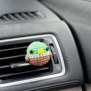 May include: A crocheted duck air freshener in a car vent. The duck has a green head, a brown body, and a yellow beak. The air vent is black, and the car's interior is dark gray.