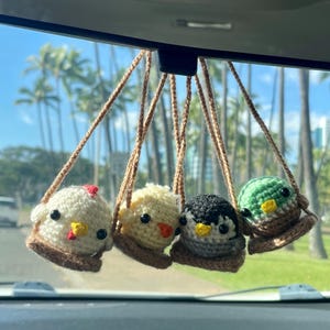 May include: Four crocheted bird ornaments hanging from a car's rearview mirror. The birds are white, yellow, black, and green, each with a different beak color. They are suspended by brown cords, with a blurred background of palm trees and a blue sky.