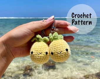 May include: Two crocheted pineapple keychains with smiling faces. The pineapples are yellow with green tops and are attached to silver key rings. The keychains are held in a hand against a blue ocean background. Crochet Pattern.