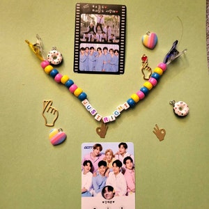 Puede incluir: Un collar de cuentas de colores con la palabra "JUST RIGHT" escrita en letras blancas. El collar tiene dijes de dona, corazón y mano. Hay dos tarjetas de fotos de una banda de chicos de K-pop, GOT7, en el fondo.