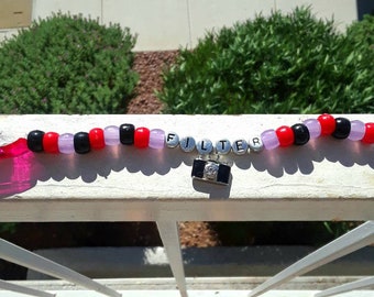 Puede incluir: Un collar de cuentas con la palabra "FILTER" deletreada con cuentas de letras plateadas. El collar presenta cuentas negras, rojas y rosas alternas. Un pequeño dije de cámara cuelga debajo de la palabra. El collar se muestra en una barandilla blanca.