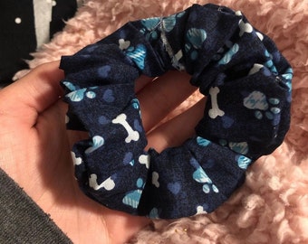 May include: A navy blue scrunchie with a pattern of white dog bones and light blue paw prints. The fabric is gathered and elasticized, creating a circular shape. This hair accessory is perfect for pet lovers.