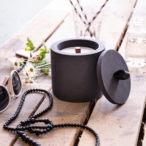 Può includere: Una candela in cemento nero con uno stoppino di legno e un coperchio. La candela è appoggiata su una superficie di legno con una collana di perline nere e occhiali da sole.