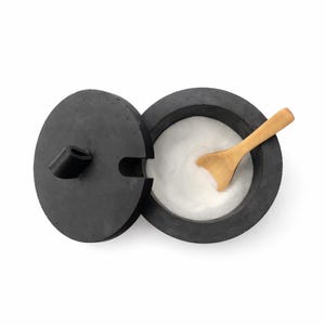 Könnte beinhalten: Ein schwarzer, runder Salzstreuer mit Deckel und einem kleinen Holzlöffel. Der Behälter ist geöffnet und zeigt weißes Salz. Der Deckel liegt daneben und zeigt das Innere des Behälters.