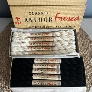 May include: Two boxes of Anchor Fresca embroidery floss. One box contains white floss, the other black. The boxes are tan with red and black text. The text on the boxes reads "CLARK'S ANCHOR Fresca Embroidery FAST COLOUR".