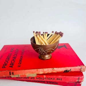 May include: A small, bronze-coloured metal bowl filled with matches, resting on a stack of red books. The bowl has a decorative etched design. The matches have yellow stems and purple tips. The books have white text.