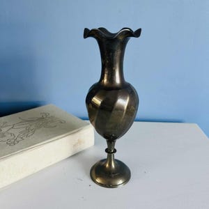 May include: A silver-coloured vase with a wavy rim and a rounded body. The vase has a pedestal base and a narrow neck. The vase is displayed on a white surface against a light blue background. A book is partially visible on the left.