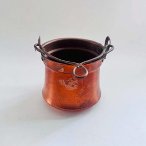 May include: A small, round copper pot with a metallic finish and a weathered patina. The vessel features a curved, hinged metal handle attached to two side loops, allowing it to swing freely. The exterior surface shows signs of oxidation.