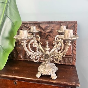 May include: A silver-toned metal candelabra featuring four candle holders arranged on ornate, curved arms. The base is wide and decorative with intricate scrollwork patterns. The structure is symmetrical, with a central finial and a textured finish.