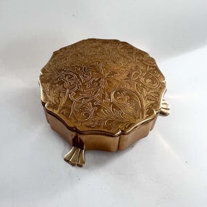 May include: A round, gold-coloured metal trinket box featuring an intricate, embossed floral and leaf pattern on the lid. The container has a scalloped edge and is supported by three small, fan-shaped feet, providing a raised, stable base.
