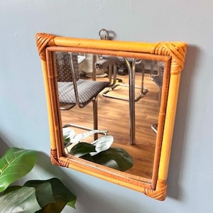 May include: A square wall mirror featuring a light brown bamboo frame with decorative woven rattan detailing at each of the four corners. The reflective glass surface shows a partial reflection of a room with chairs and green plant leaves.