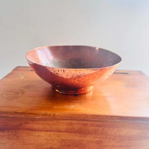 May include: A round, hammered copper bowl with a metallic reddish-brown finish. The bowl features a wide, open top and a small, circular base, showcasing a textured surface created by individual hammer marks across the exterior of the metal.