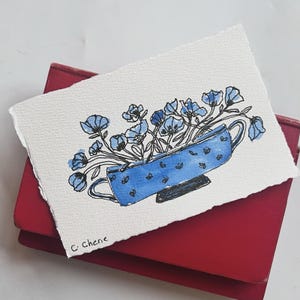 May include: Watercolour painting of blue flowers in a blue teacup with a handle, set against a white background. The teacup has a pattern of small, dark blue shapes. The artwork is signed "C. Cherie" in the lower left corner. The painting rests on a red surface.
