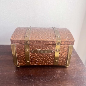 May include: A rectangular tea chest with a domed lid, featuring a textured copper-coloured surface accented by polished brass vertical bands and corner reinforcements. A brass plaque on the front is engraved with the word TEA in capital letters.