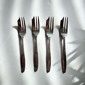 May include: A set of four silver-toned metal forks arranged in a vertical row. Each fork features three tines, with the left tine being wider and pointed, and an ornate, embossed floral pattern along the handle stem leading to a smooth base.