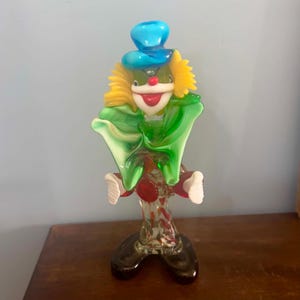 May include: A glass clown figurine featuring a bright blue hat, yellow hair, and a green ruffled collar. The clown has a white face with a red nose and mouth, a clear body with red and white accents, and large dark brown shoes at the base.