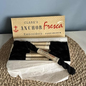 May include: A box of black Anchor Fresca embroidery thread. The box is tan with red text that reads "Clark's Anchor Fresca Embroidery Fast Colour." The box contains several spools of black thread, with one spool unwound.
