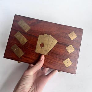 May include: A rectangular wooden box with brass inlays of playing cards, dominoes, and dice. The box is a rich brown colour, and the brass inlays are a warm gold. The box is held by a hand.