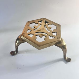 May include: A hexagonal brass trivet featuring an openwork scroll pattern and three curved legs. The metal surface has a polished gold-toned finish with visible reflections. The design includes symmetrical cutouts throughout the top plate.