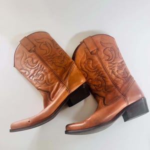 May include: A pair of tan leather cowboy boots featuring a square toe, a stacked black heel, and decorative stitching patterns on the shaft. Each boot includes pull tabs at the top and exhibits a smooth finish with subtle tonal variations.