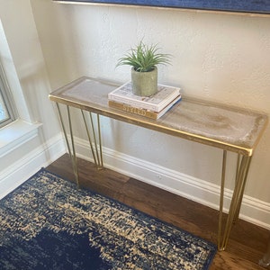 May include: A gold metal console table with a white top and three hairpin legs. The table is decorated with two books and a potted plant. The table is sitting on a blue and beige patterned rug.