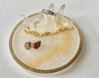 May include: A white and gold ring dish with a cluster of clear quartz crystals in the center. Two small gold earrings are on the dish.