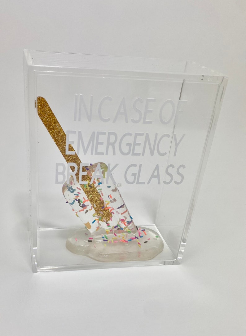 May include: A clear acrylic box containing a gold glitter popsicle. The popsicle is melting and has colourful sprinkles on it. The text on the box reads "In case of emergency break glass."