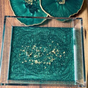May include: A rectangular, clear acrylic tray with a deep emerald green resin base and gold leaf accents. Two matching coasters with gold edges are visible in the background. The tray is on a wooden surface.