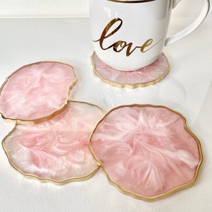 Peut inclure: Quatre sous-verres en résine rose et blanche avec une bordure dorée. Les sous-verres ont la forme d'un cercle libre. Une tasse blanche avec une bordure dorée et le mot "love" écrit en or est posée sur l'un des sous-verres.