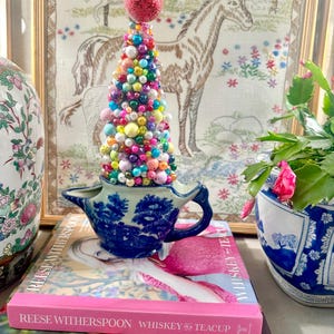 May include: A whimsical Christmas tree made of colorful beads sits atop a blue and white porcelain creamer. The tree is topped with a red glittery ball. The creamer rests on a stack of books, including "Reese Witherspoon Whiskey Teacup."
