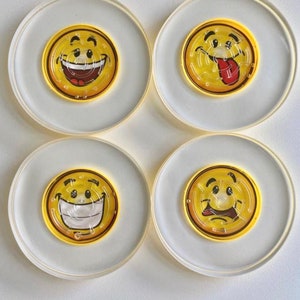 May include: Four clear acrylic coasters with yellow smiley face mazes in the center. Each coaster features a different expression: laughing, sticking out tongue, smiling, and sad. The coasters are round with a white background.