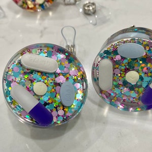 May include: Clear, circular ornaments filled with colorful glitter and various pills. One ornament has a white and blue capsule, a white pill, a light blue pill with "1363" on it, and a small yellow pill. The other ornament has a white pill and a light blue pill.