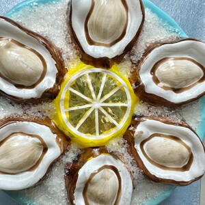 May include: A ceramic oyster serving platter with six oyster shells arranged around a yellow lemon slice. The oysters have brown and white shells, and the platter is light blue with a textured surface.
