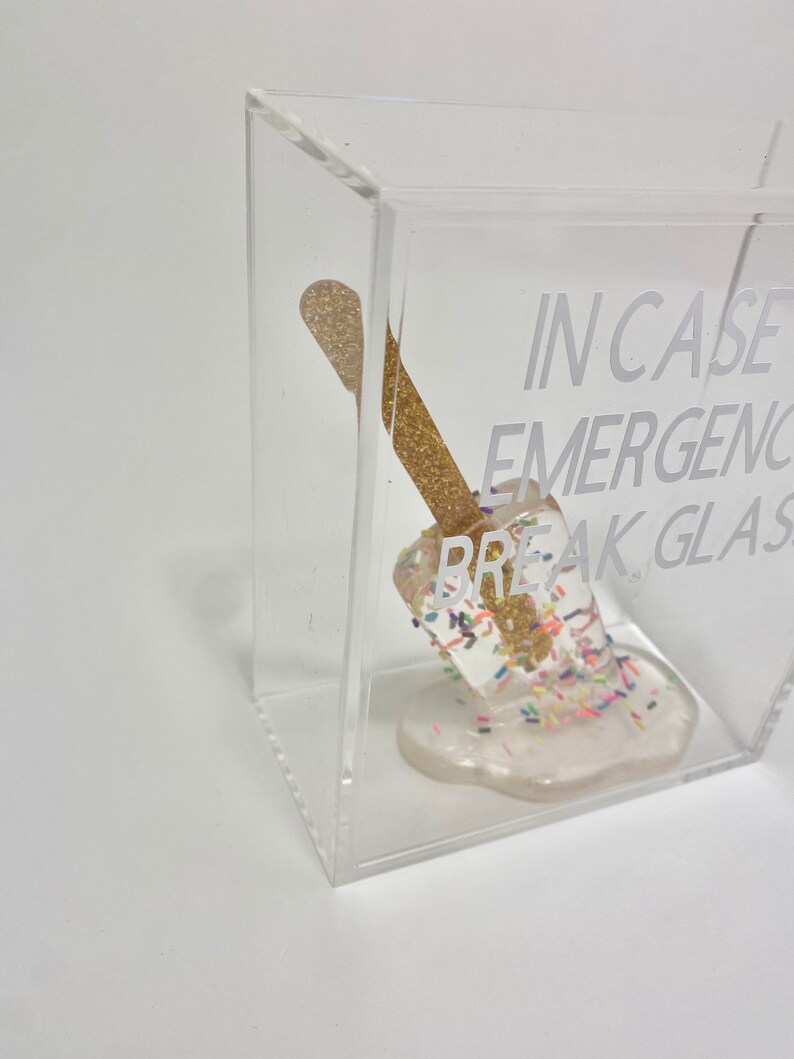 May include: A clear acrylic box containing a gold and white popsicle. The popsicle is melting and has sprinkles on it. The text "IN CASE OF EMERGENCY BREAK GLASS" is printed on the box.