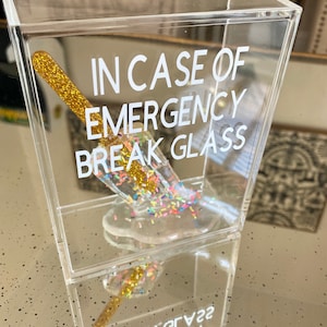 May include: A clear acrylic box containing a gold glitter stick and a clear glass filled with colourful sprinkles. The box has the text "IN CASE OF EMERGENCY BREAK GLASS" printed on the front.
