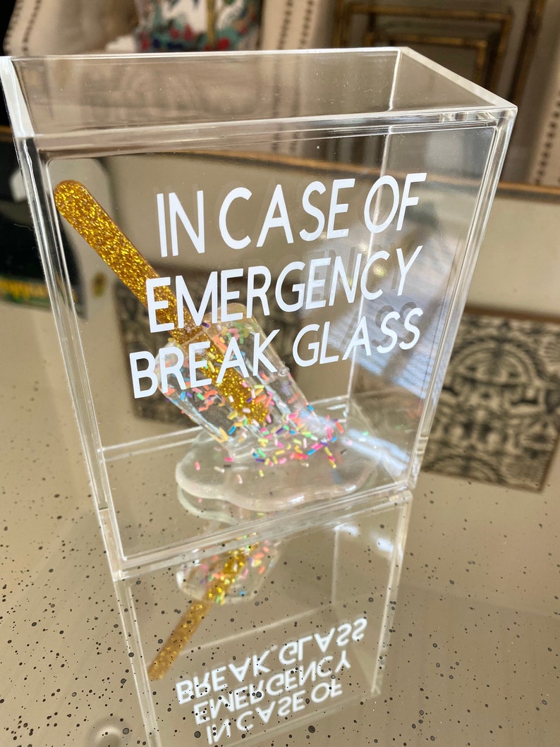 May include: A clear acrylic box containing a gold glitter stick and a clear glass filled with colourful sprinkles. The box has the text "IN CASE OF EMERGENCY BREAK GLASS" printed on the front.