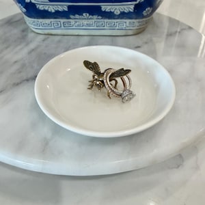 May include: A small white ceramic trinket dish featuring a metallic bronze-toned bee figurine in the centre. Two silver-toned rings with clear stones are resting on the back of the bee, which is positioned inside the circular bowl.