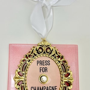 Puede incluir: Un adorno rectangular rosa con un marco ovalado dorado y ornamentado. El marco rodea un óvalo rosa con las palabras "PRESS FOR CHAMPAGNE" y un botón perlado. Se adjunta una cinta blanca para colgar.