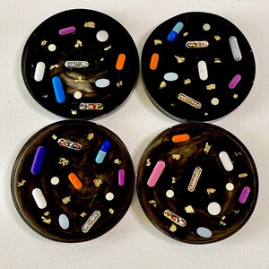 May include: Four black round coasters with a white, gray, and black marbled design. Each coaster is decorated with colorful pills and gold flakes.
