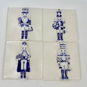 May include: Set of four square coasters featuring blue and white nutcracker soldier designs. Each coaster has a different nutcracker illustration, with details like a hat, uniform, and accessories. The coasters are on a white background.