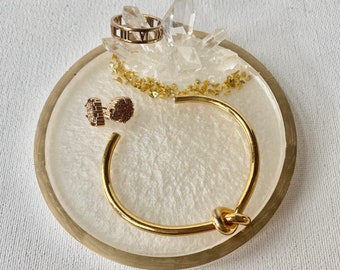 May include: A white resin ring dish with a gold rim. The dish features a clear quartz crystal cluster and gold glitter. A gold bangle with a knot detail, a gold ring with Roman numeral markings, and two gold earrings are displayed on the dish.