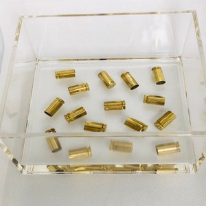 May include: A clear acrylic tray filled with several brass bullet casings. The casings are scattered inside the tray, which sits on a white surface. The casings are a shiny gold color.