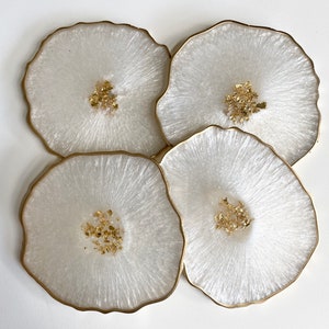 May include: Four white coasters with gold flecks and a gold rim. The coasters are round and have a smooth, glossy finish.