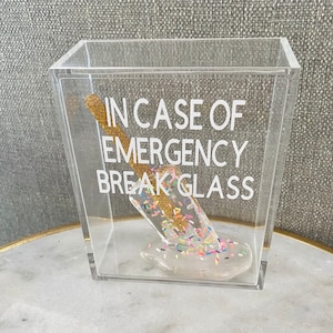 May include: A clear acrylic box containing a gold glitter stick and a white melted ice cream cone with sprinkles. The box has the text "In case of emergency break glass" printed on the side.