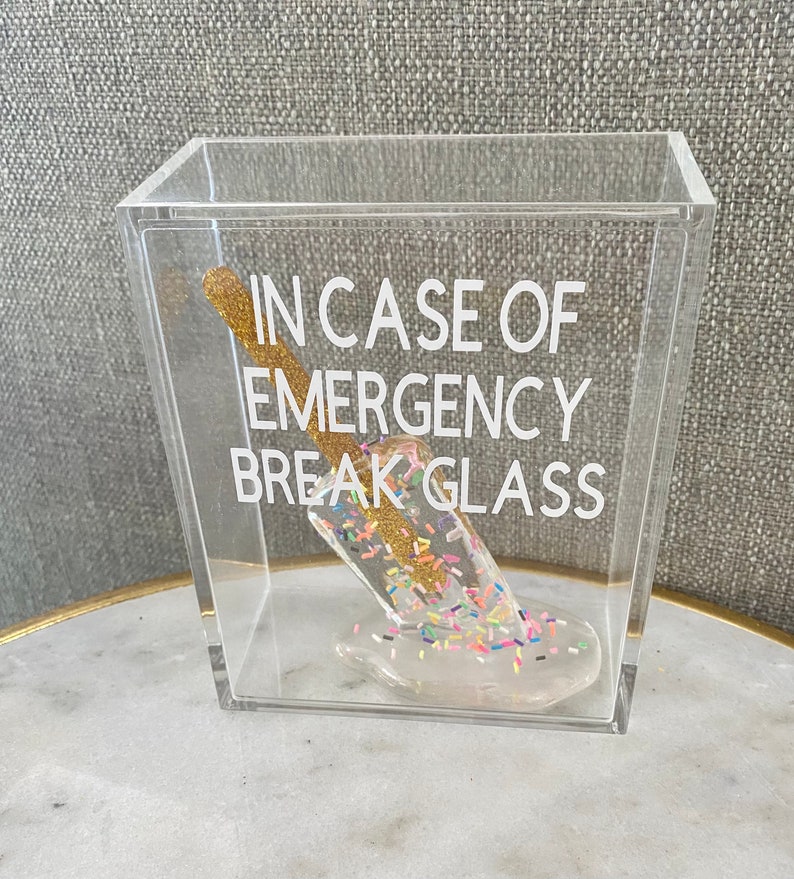 May include: A clear acrylic box containing a gold glitter stick and a white melted ice cream cone with sprinkles. The box has the text "In case of emergency break glass" printed on the side.