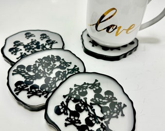 Peut inclure: Ensemble de quatre sous-verres en forme de crâne et d'os croisés, blancs et noirs. Les sous-verres ont une bordure noire et un fond blanc avec des motifs de crânes et d'os croisés noirs. Les sous-verres sont sur une surface blanche.