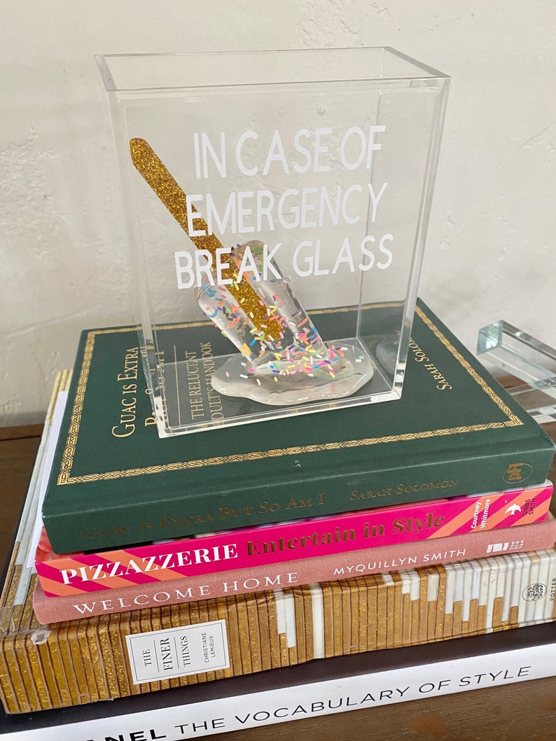 May include: A clear acrylic box containing a gold glitter popsicle. The box has the text "In case of emergency break glass" printed on the front. The box is sitting on top of a stack of books.