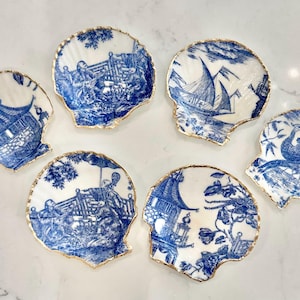 May include: Set of six seashell-shaped decorative dishes with blue and white Chinoiserie designs. Each dish features a different scene, with gold-painted edges. The dishes are arranged on a white surface.
